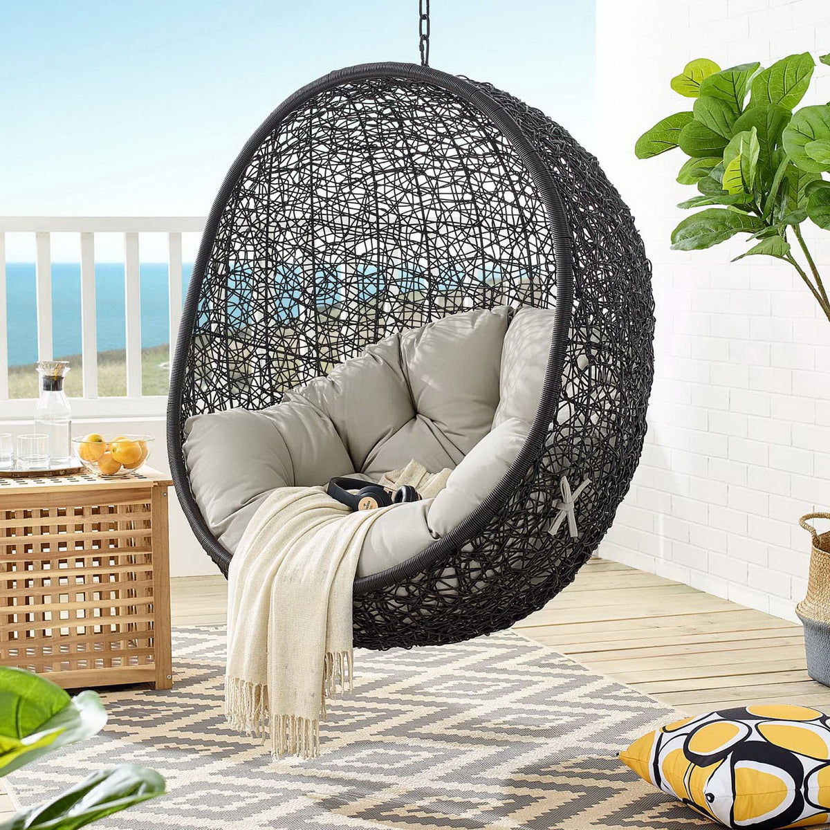 Beige cushion swing chair hangs on a breezy deck with ocean view and patterned rug || EEI-739-BEI-SET || front_partial_room