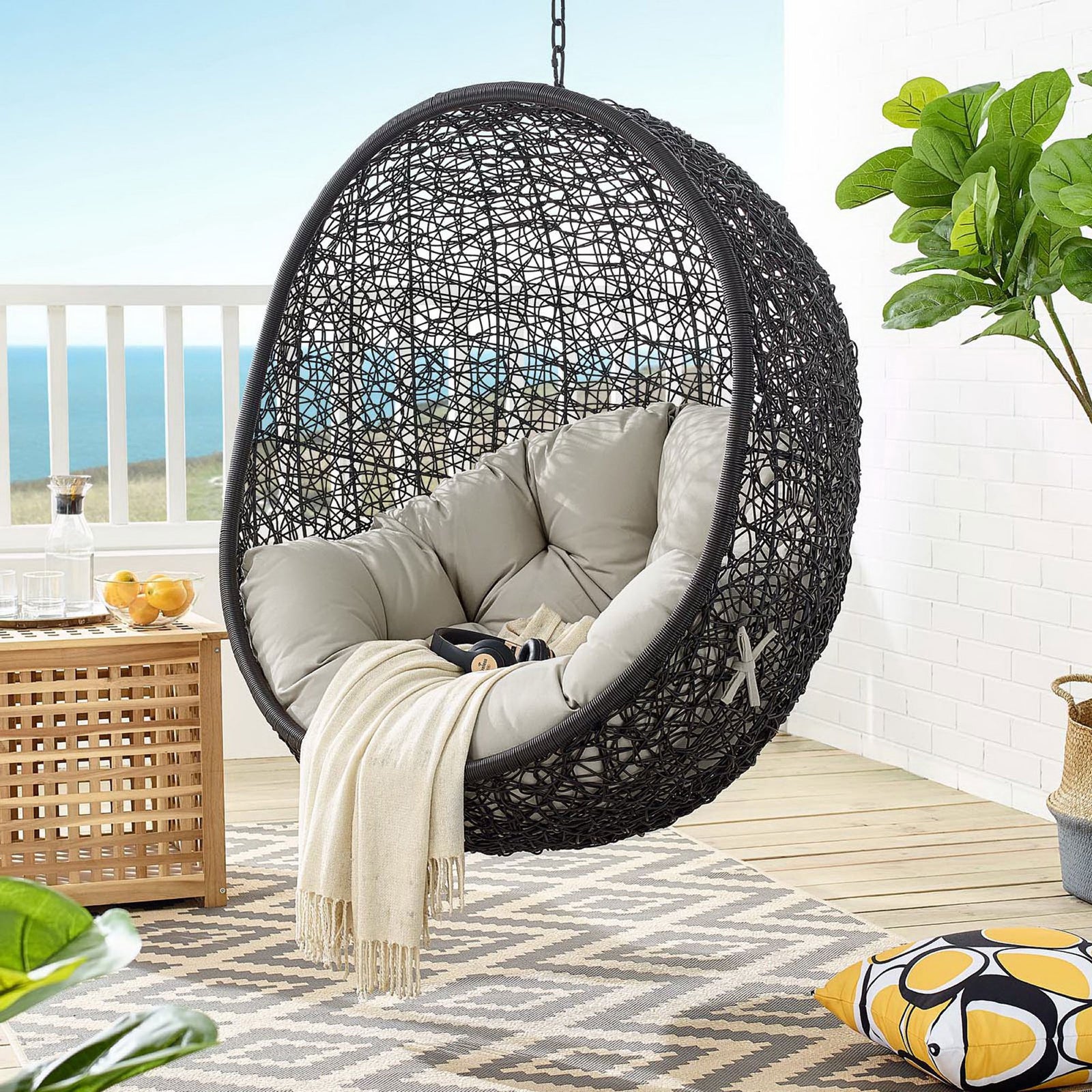 Beige cushion swing chair hangs on a breezy deck with ocean view and patterned rug || EEI-739-BEI-SET || front_partial_room