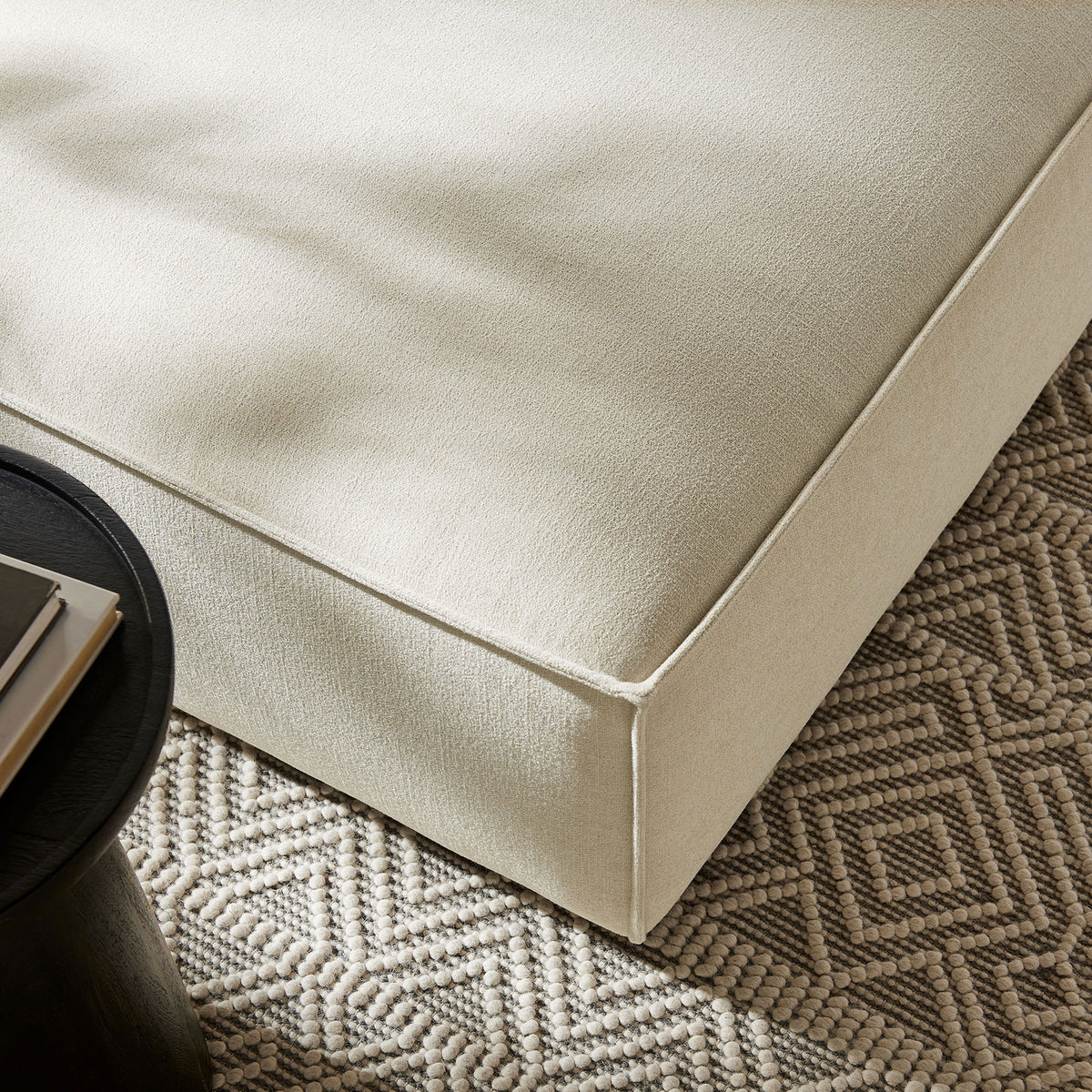 Detail of ecru ottoman upholstery styled with textured area rug and modern black accent table. || EEI-7401-ECR || detail_room