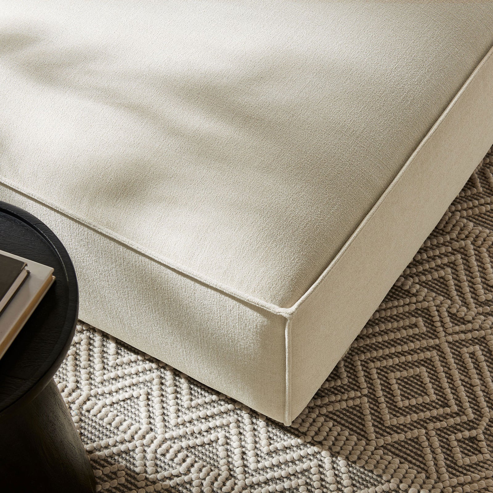 Detail of ecru ottoman upholstery styled with textured area rug and modern black accent table. || EEI-7401-ECR || detail_room