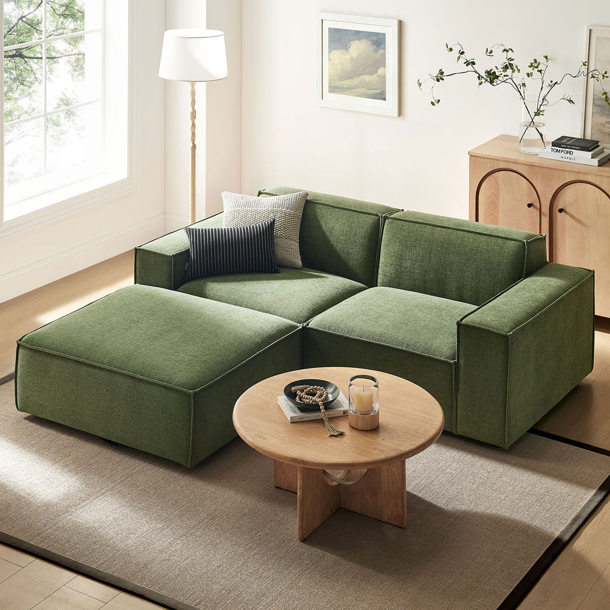 Green modular loveseat with ottoman anchoring a bright living room, paired with a round wood coffee table || EEI-7405-FOL || front_angle_room