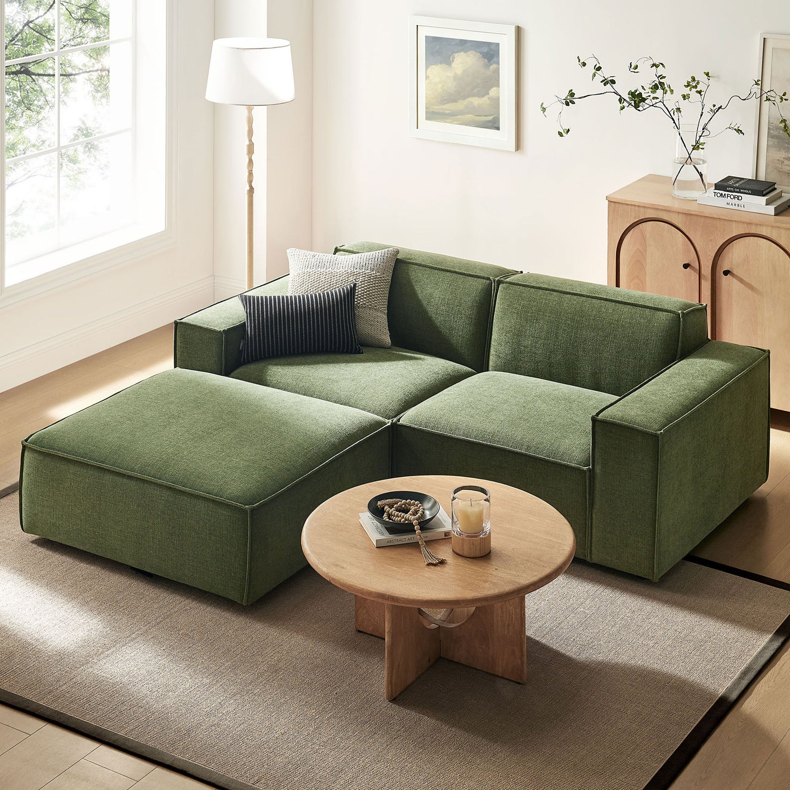 Green modular loveseat with ottoman anchoring a bright living room, paired with a round wood coffee table || EEI-7405-FOL || front_angle_room