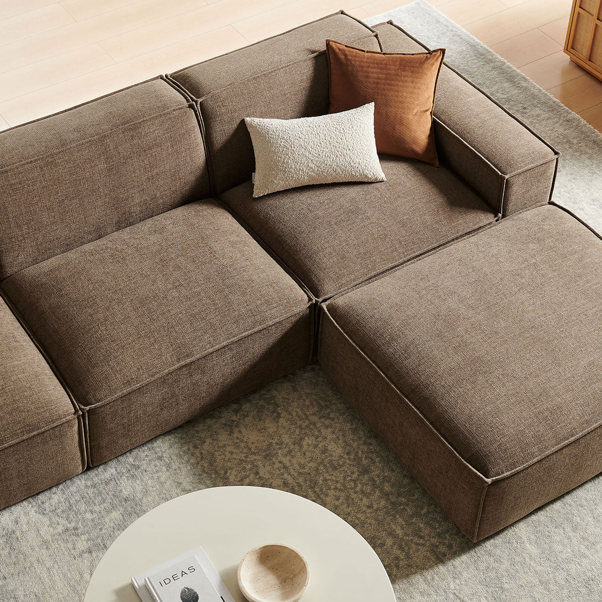 Cozy corner of the mocha modular sofa with textured pillows and ottoman over a soft neutral rug. || EEI-7409-MOC || detail_room