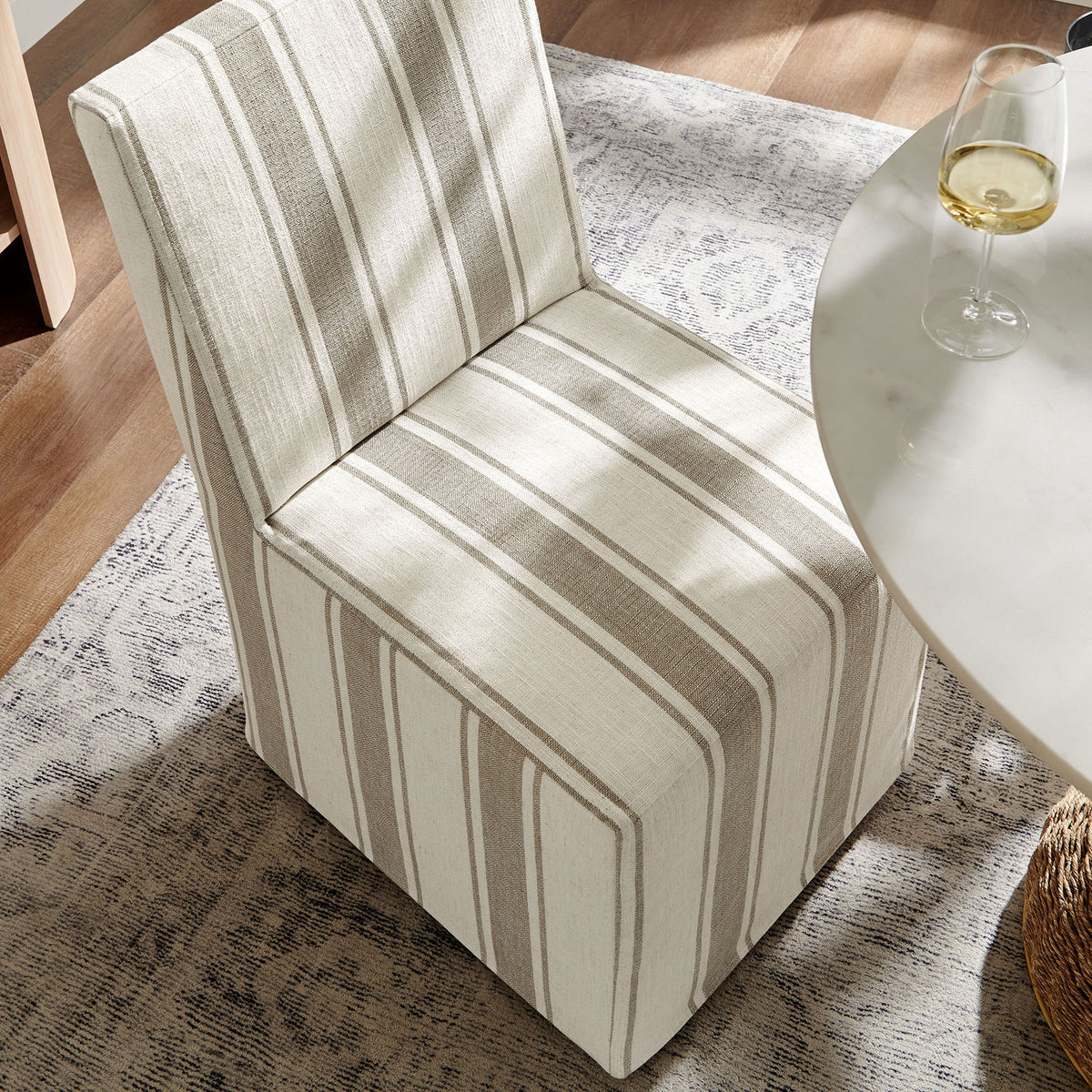 Tan striped slipcovered dining chair gliding easily beside a round table on a soft patterned area rug || EEI-7486-TAN || front_angle_room