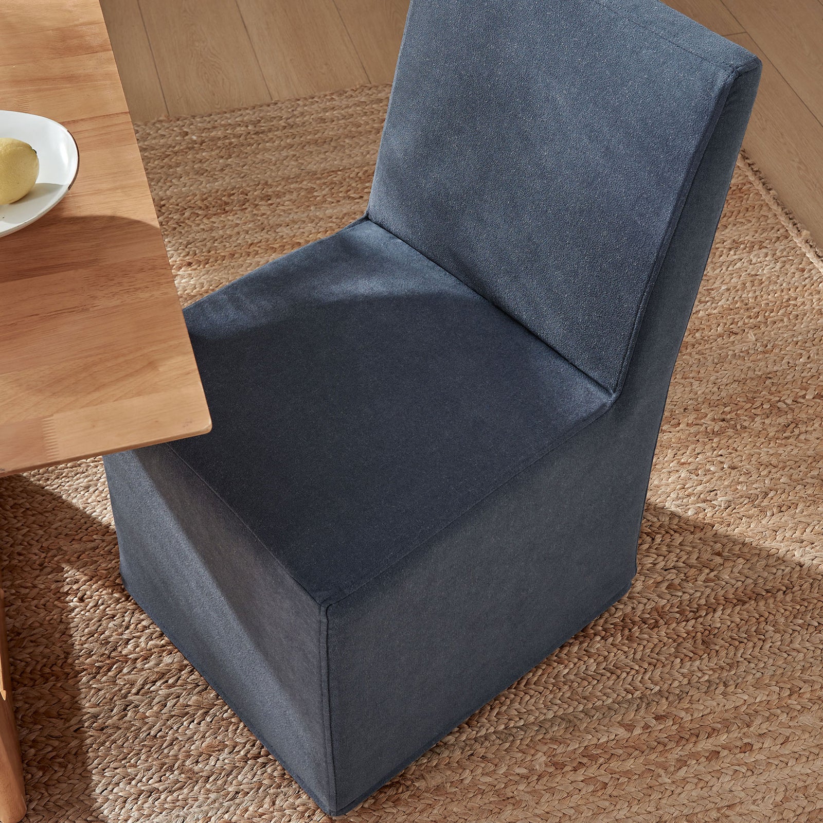 Denim slipcovered dining chair tucked beside a light wood table on a woven natural rug || EEI-7528-DEN || front_angle_room