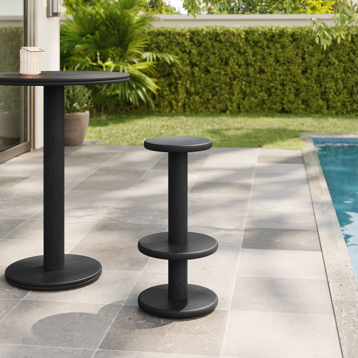 Black acacia bar stool and pedestal bar table on a poolside patio with lush greenery || EEI-7586-BLK || front_angle_room