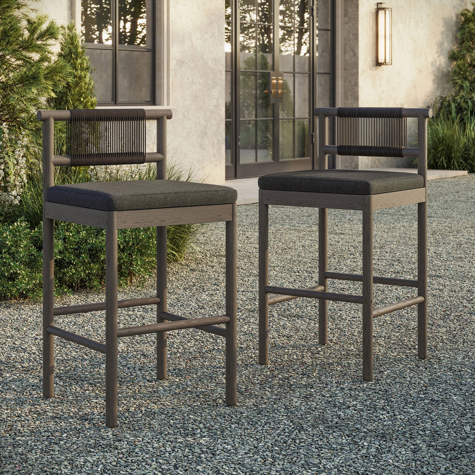 Pair of weathered gray bar stools arranged at an outdoor bar table on a gravel terrace || EEI-7626-WGR-FLI || front_angle_multi_room