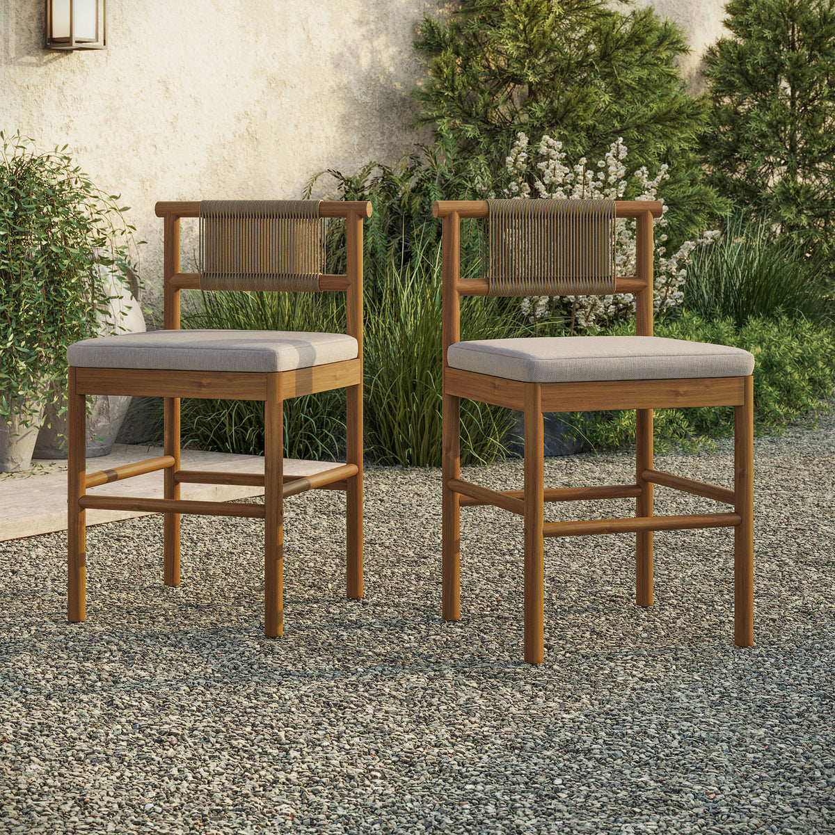 Pair of teak counter stools arranged at an outdoor bar table on a gravel patio || EEI-7627-NAT-SAI || front_angle_multi_room