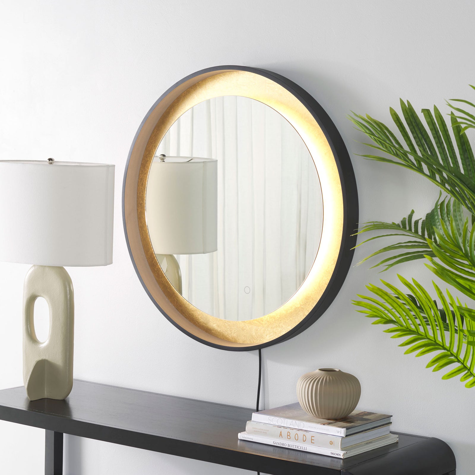 A warm LED glow illuminates the gold-leaf surround of a black-framed mirror above a console. || LMR1014B || front_angle_on_room