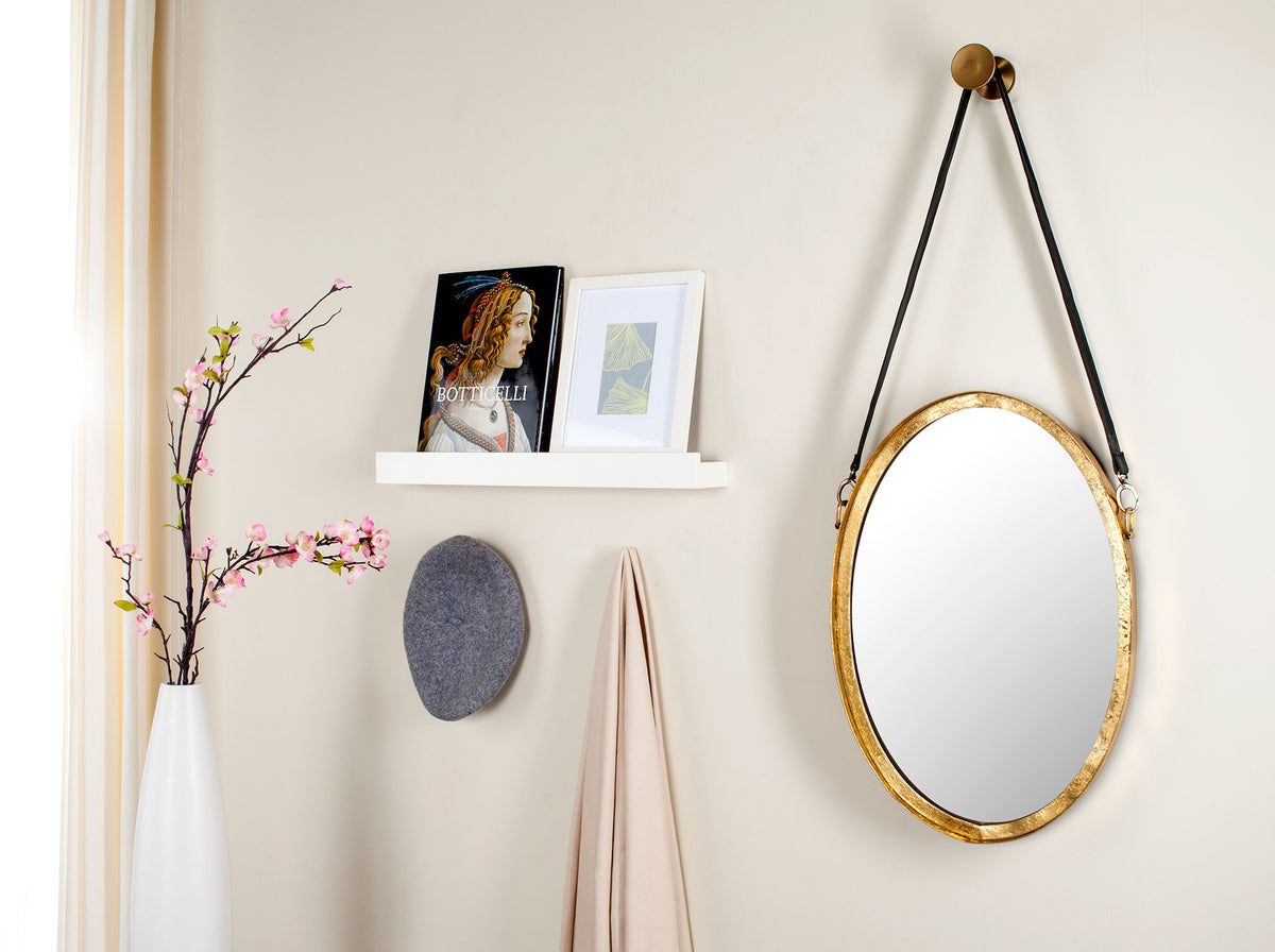 A gold-framed oval mirror with a black hanging strap brings warmth and depth to a bright wall.