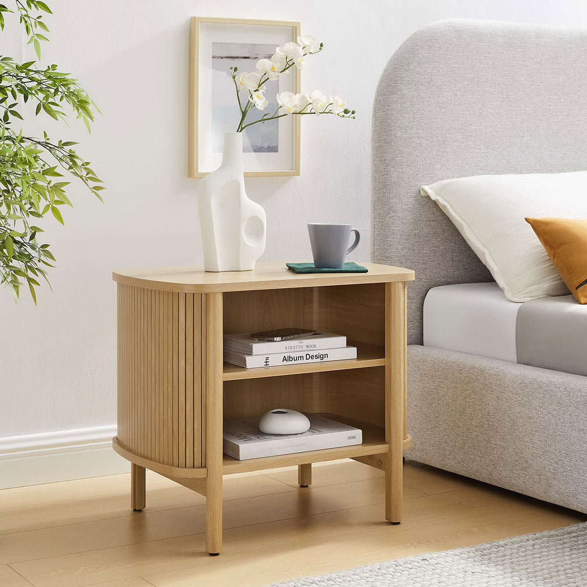 Cadence oak nightstand beside a modern upholstered bed, styled with decor to showcase its two open storage shelves || MOD-7157-OAK || front_angle_room