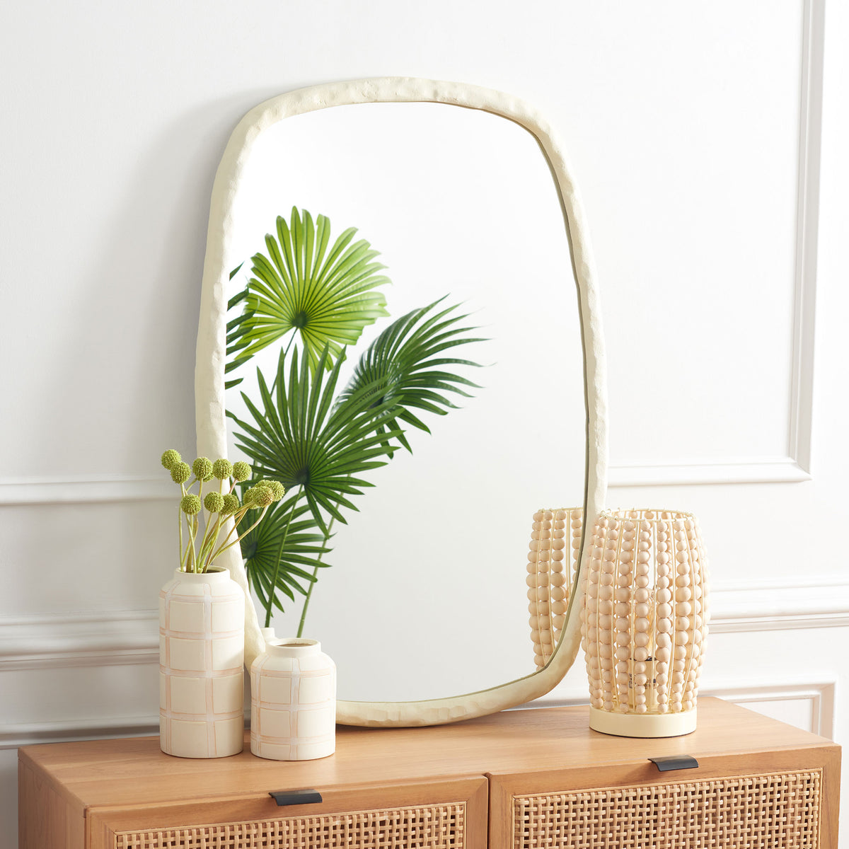 Sculpted ivory mirror brings soft curves above a wood cabinet and reflects lush greenery. || MRR3068A || front_room