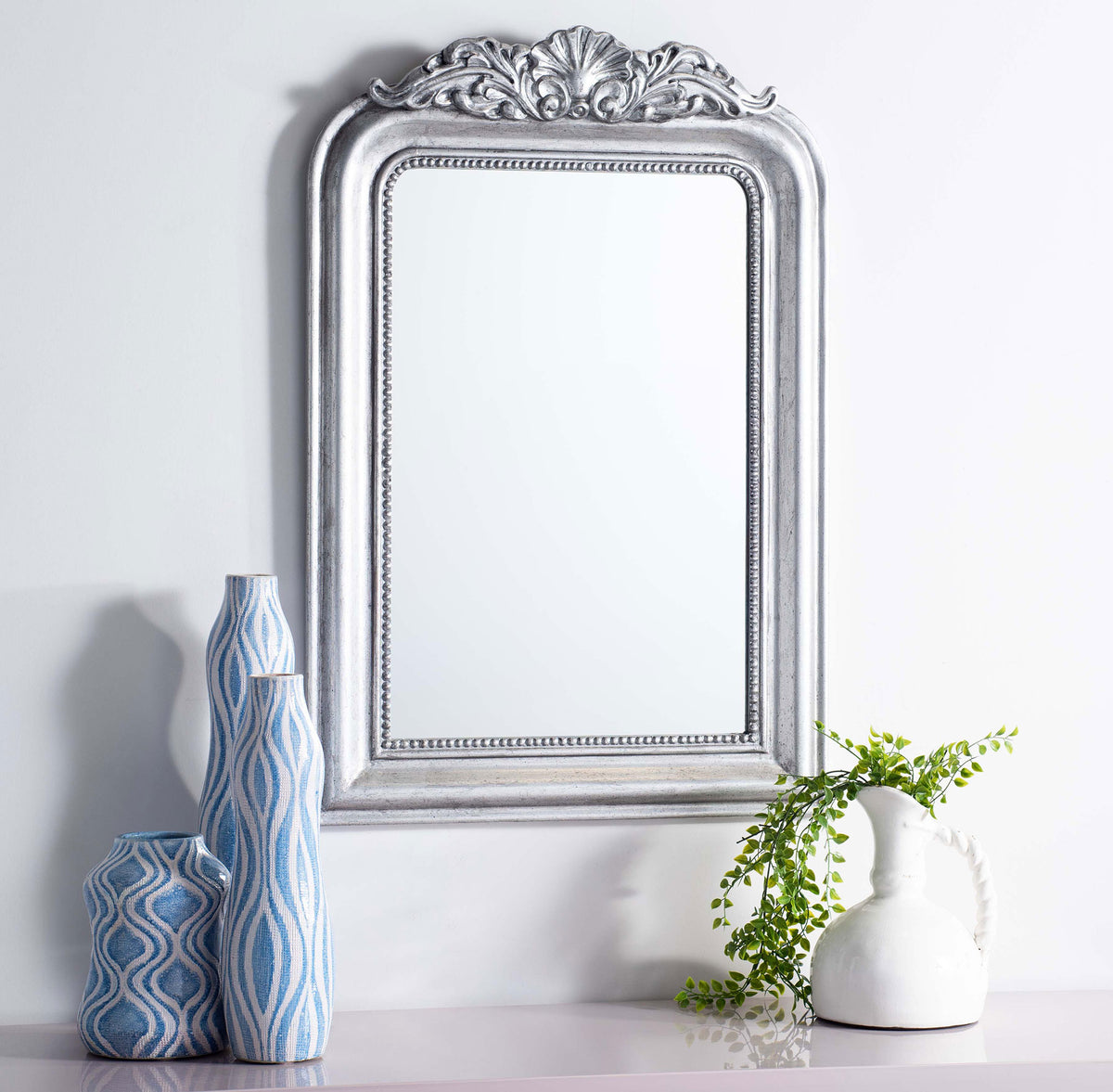 Antique silver mirror brings ornate detail to a wall above blue vases and greenery