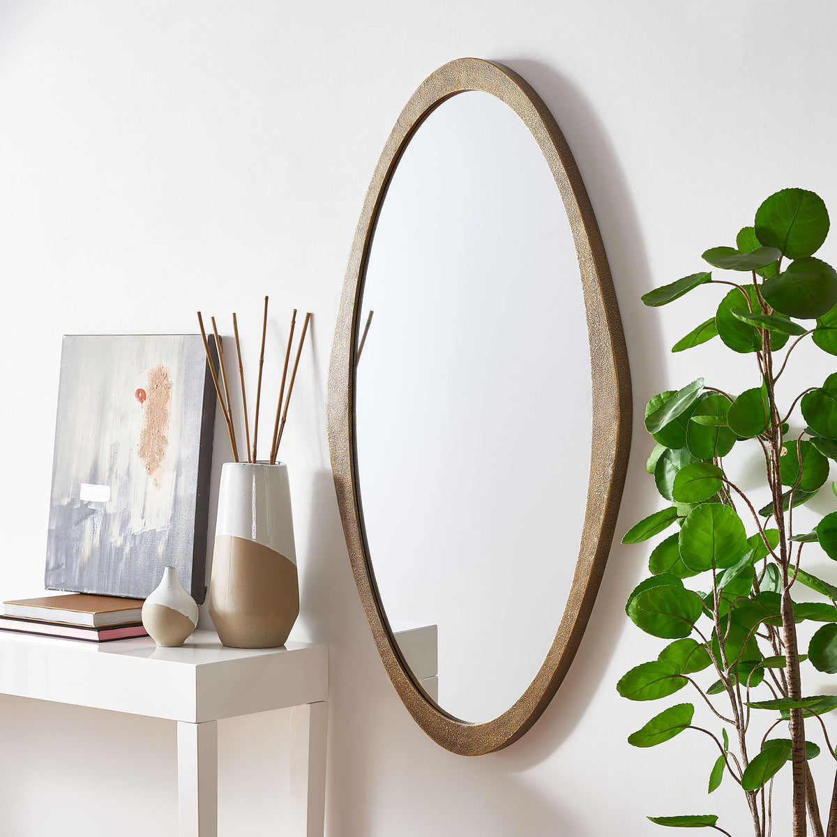 A brass-framed oval mirror adds warm metallic character beside a white console and greenery.