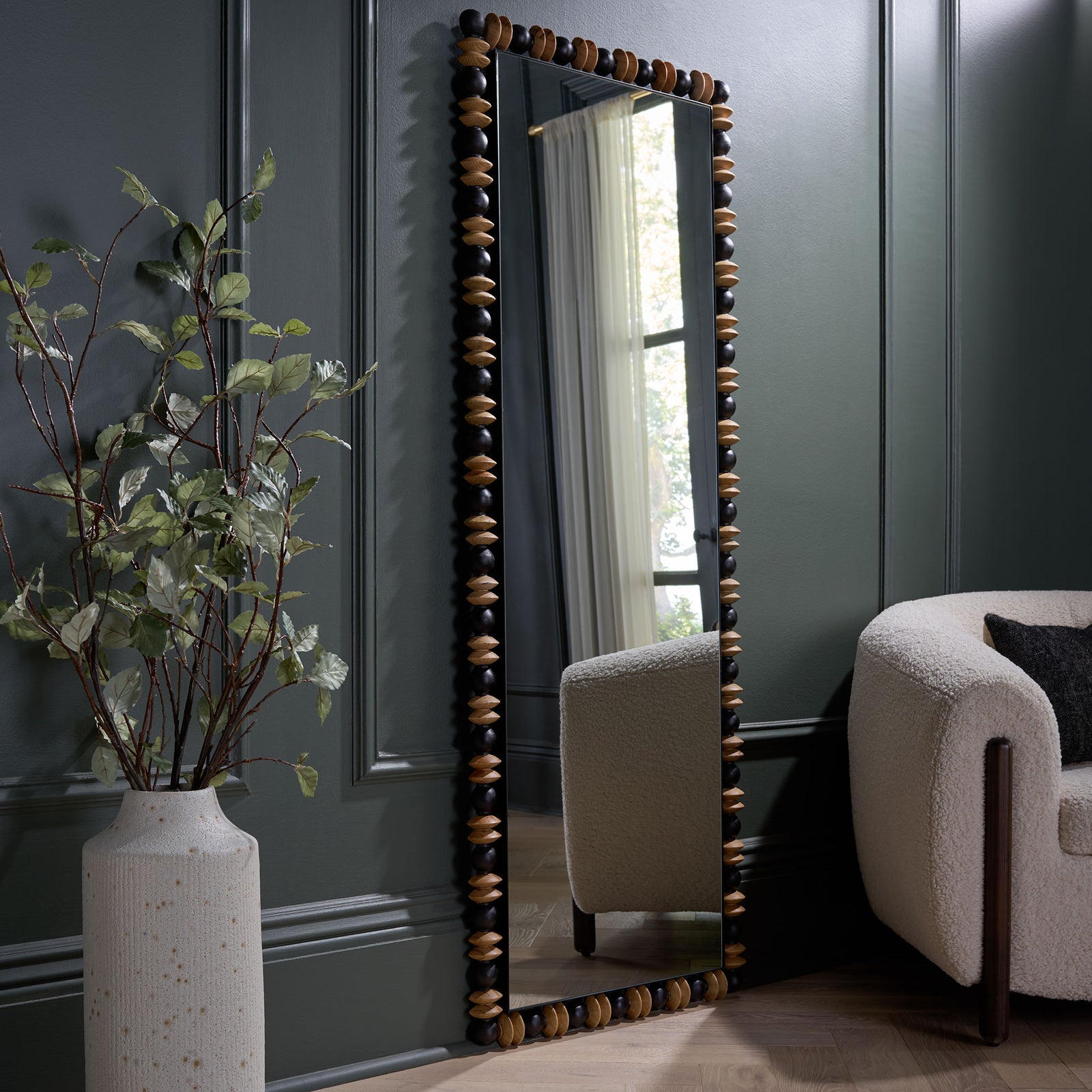 A tall mirror with a black and natural wood border adds sculptural warmth against a dark paneled wall. || MRR9028A || front_angle_room