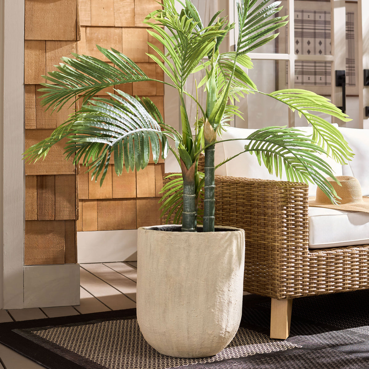 A leafy palm rises from the compact beige planter beside woven patio seating. || PAT1404B || front_room