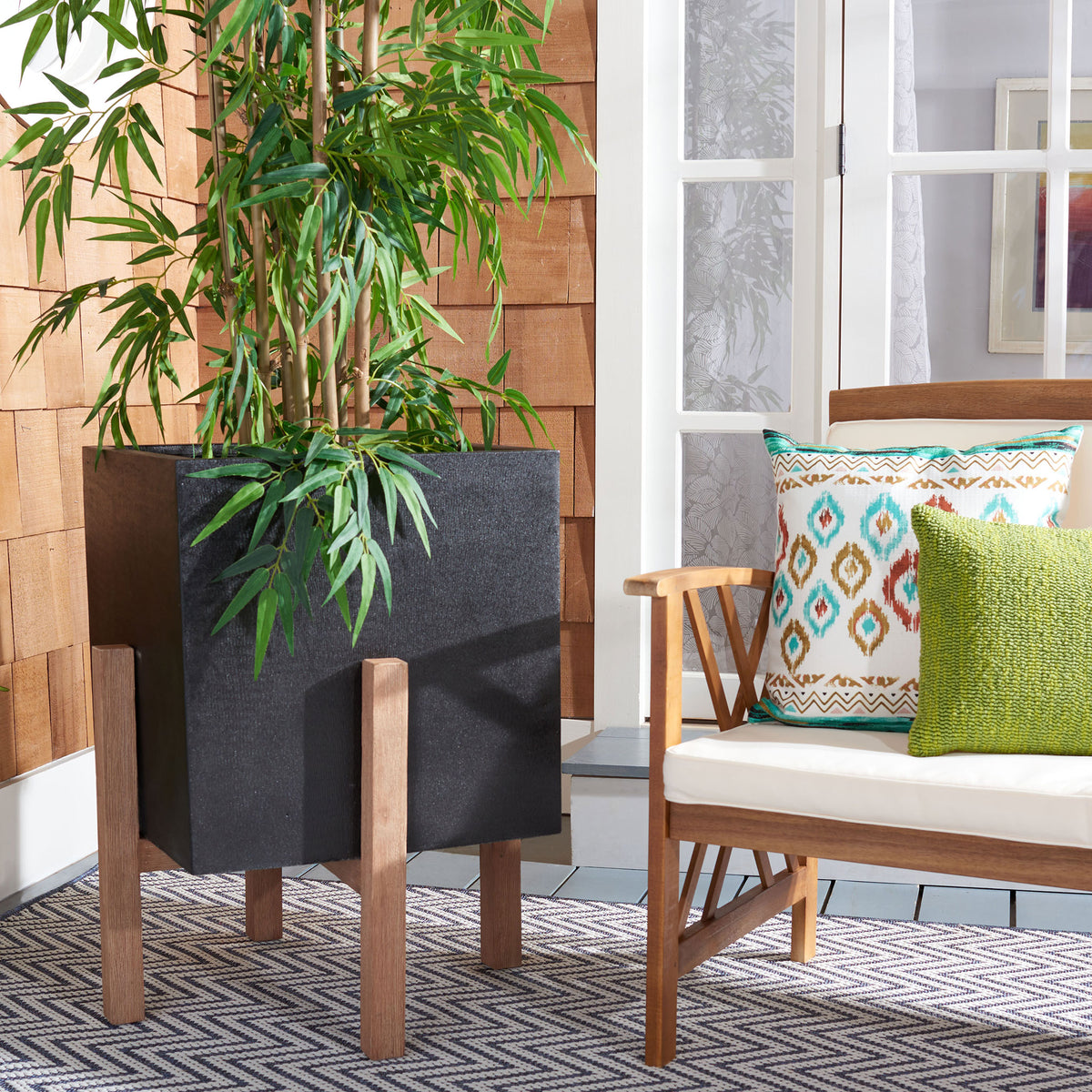 A lush leafy plant rises from the large black planter beside a cushioned wooden bench. || PAT1513A || front_angle_room