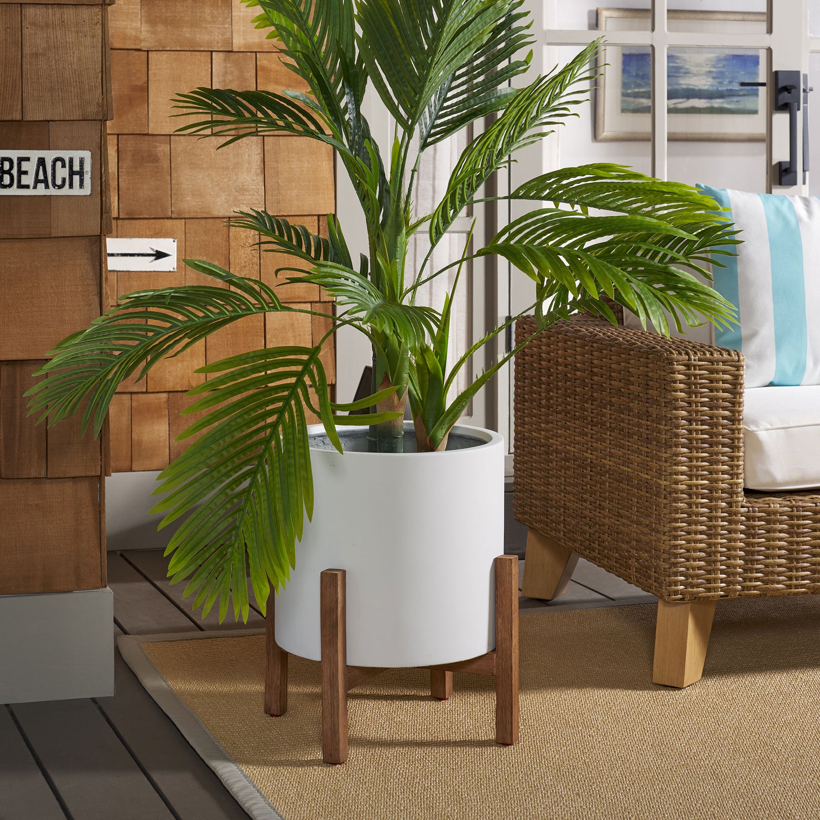 Leafy palms fill a white planter elevated on wood-grain legs alongside a woven chair. || PAT1514C || front_room