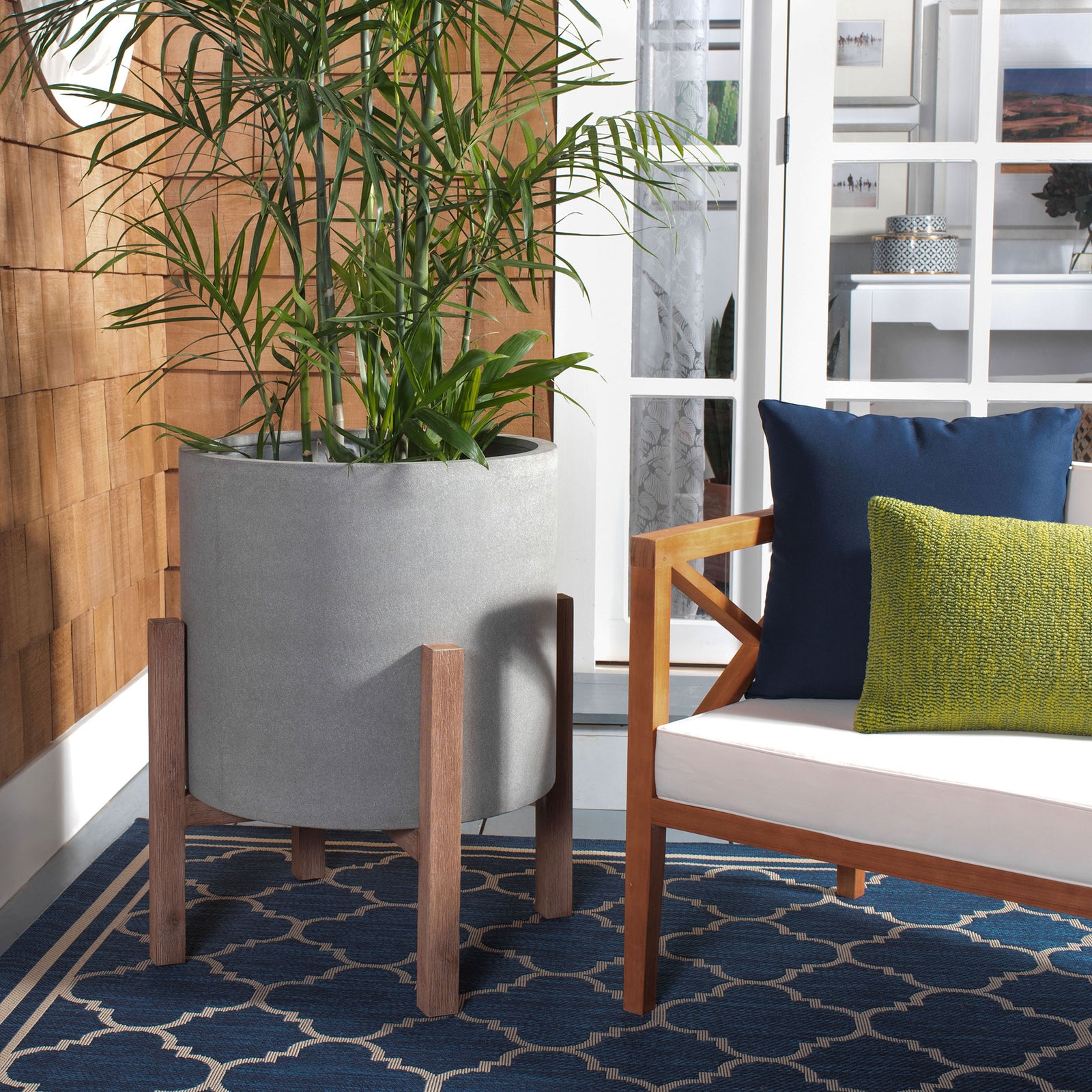 Tall tropical foliage fills a gray planter elevated on wood-toned legs by a cushioned chair. || PAT1515B || front_room