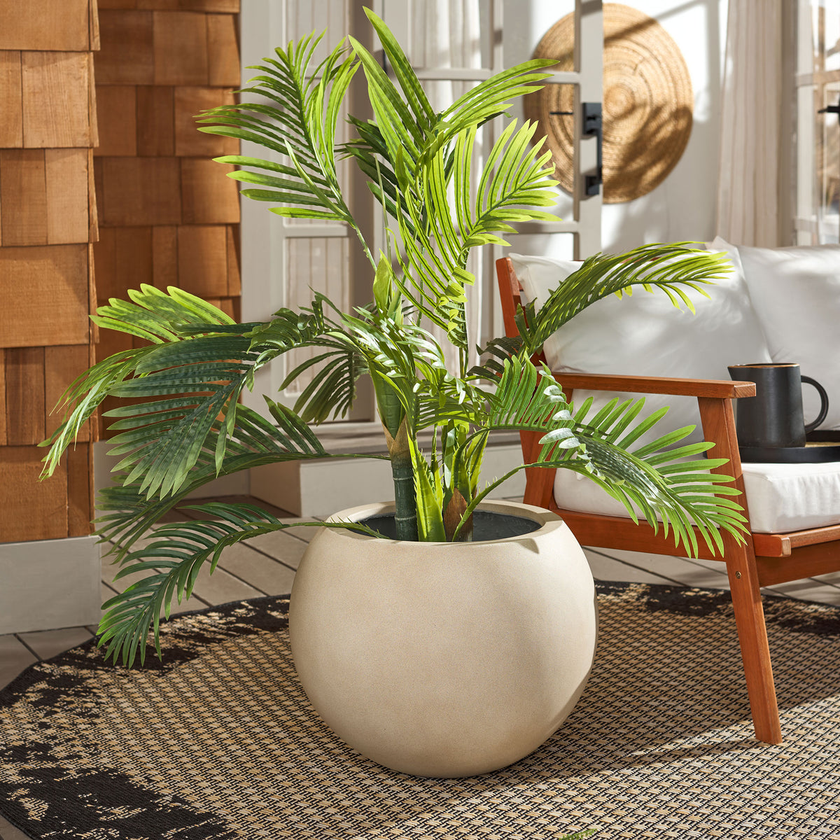 A large beige orb planter holds lush palm fronds alongside warm wood seating. || PAT1519B || front_room