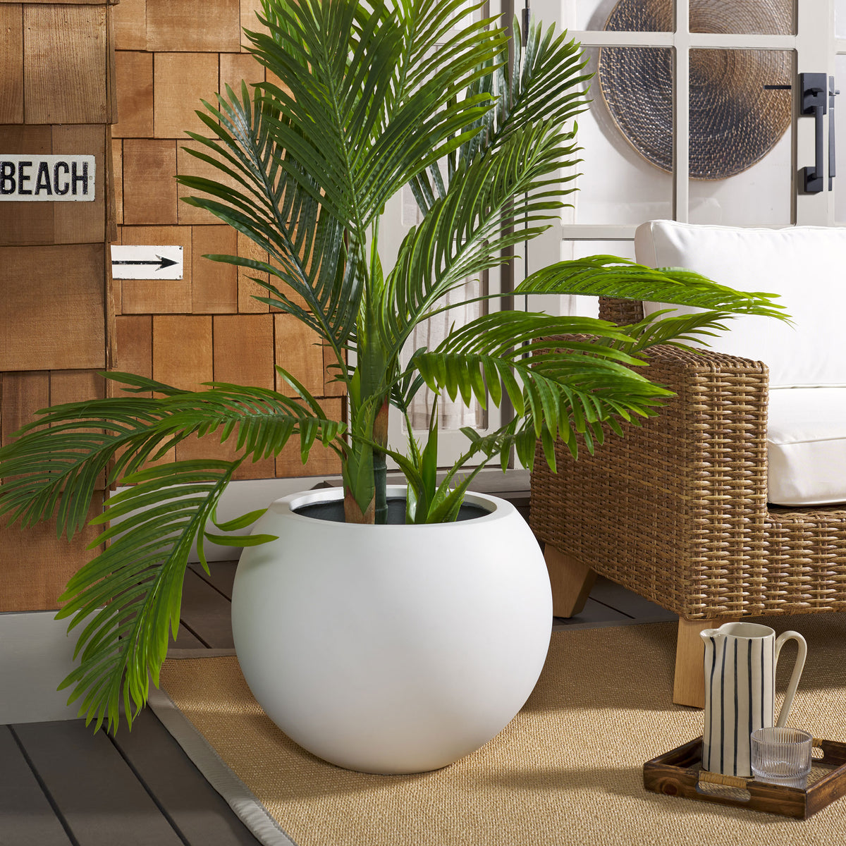 A broad white planter anchors tropical greenery beside wicker seating and a woven rug. || PAT1519C || front_room