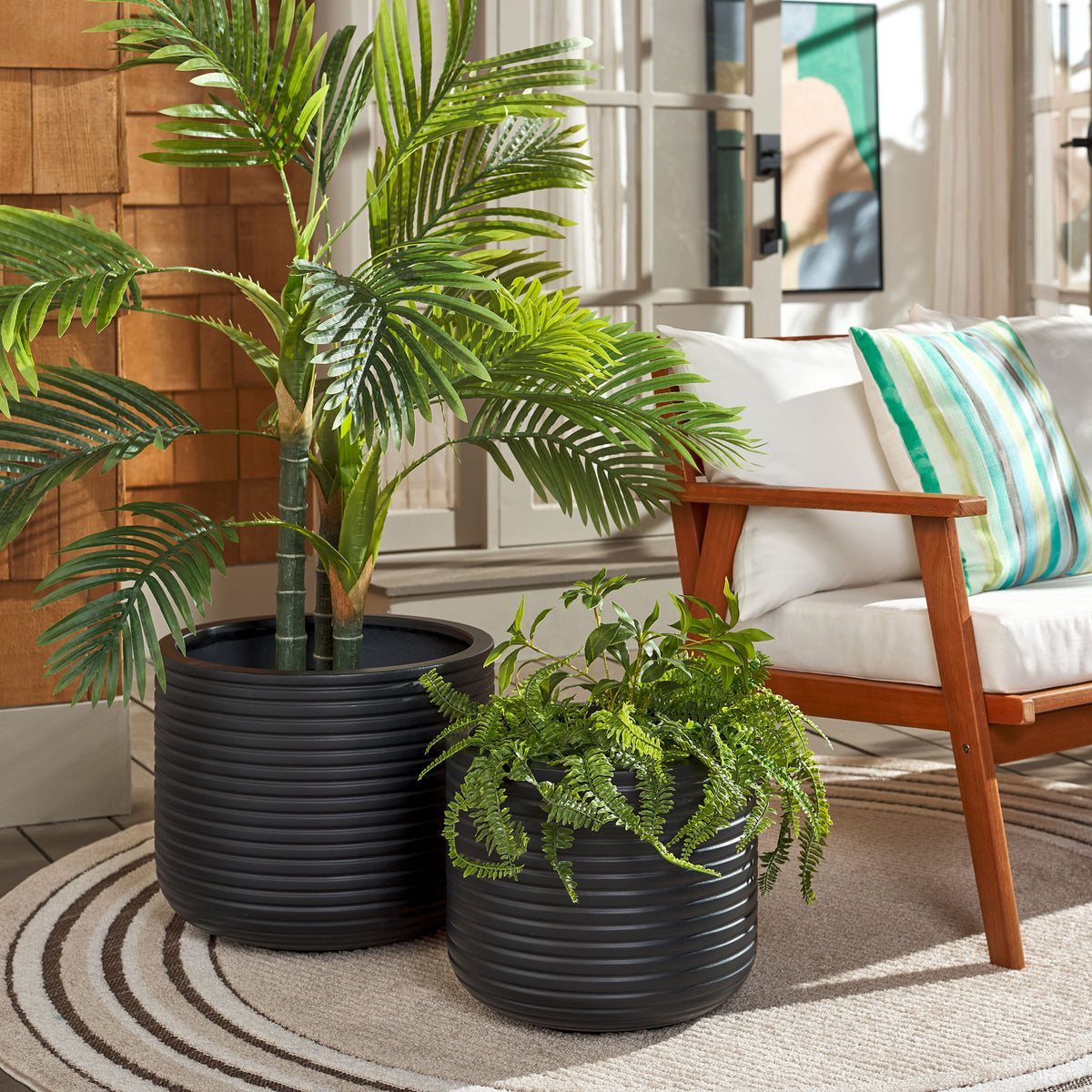 Two black ribbed planters frame lush greenery beside a cushioned wood-framed chair