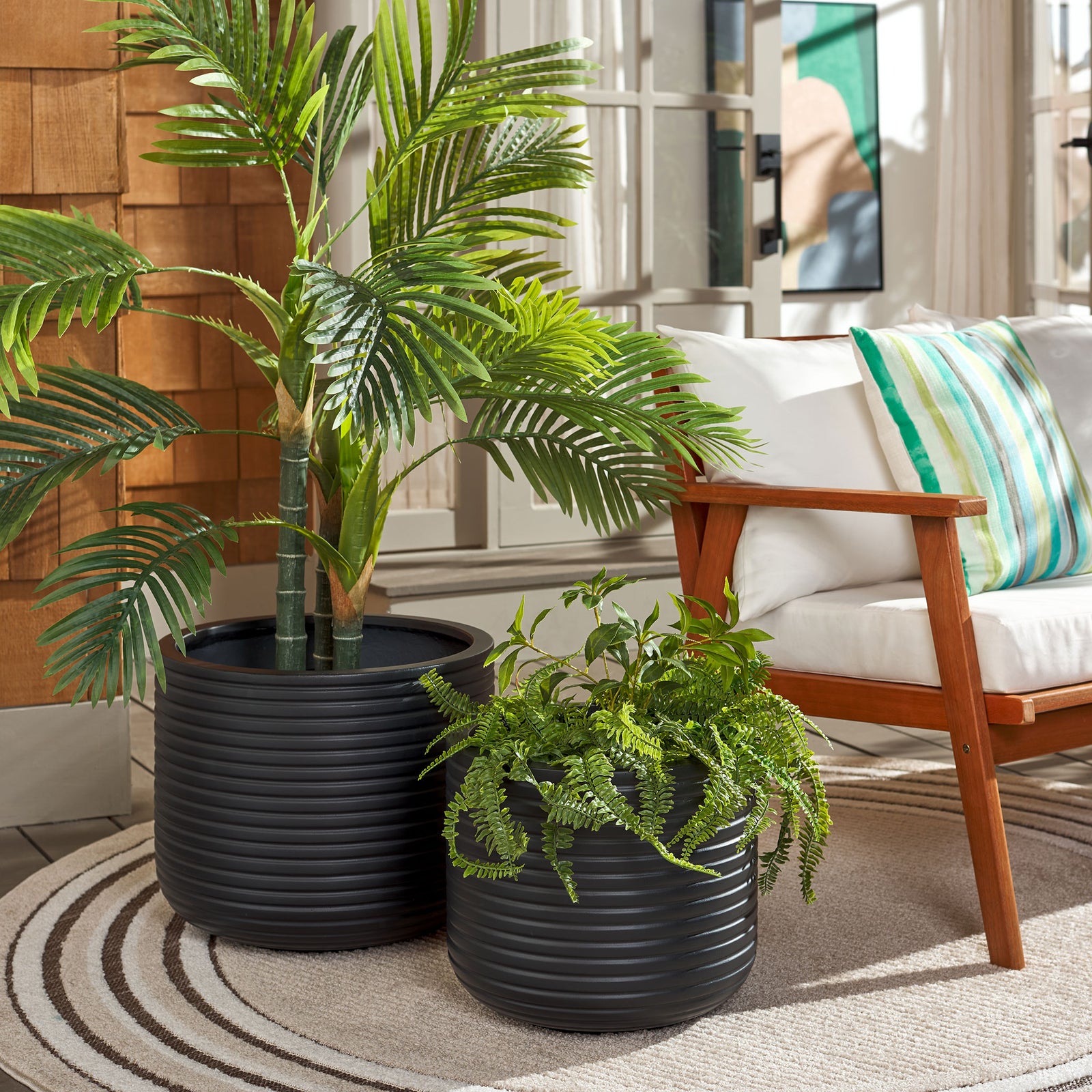 Two black ribbed planters frame lush greenery beside a cushioned wood-framed chair || PAT1530A-SET2 || front_angle_room