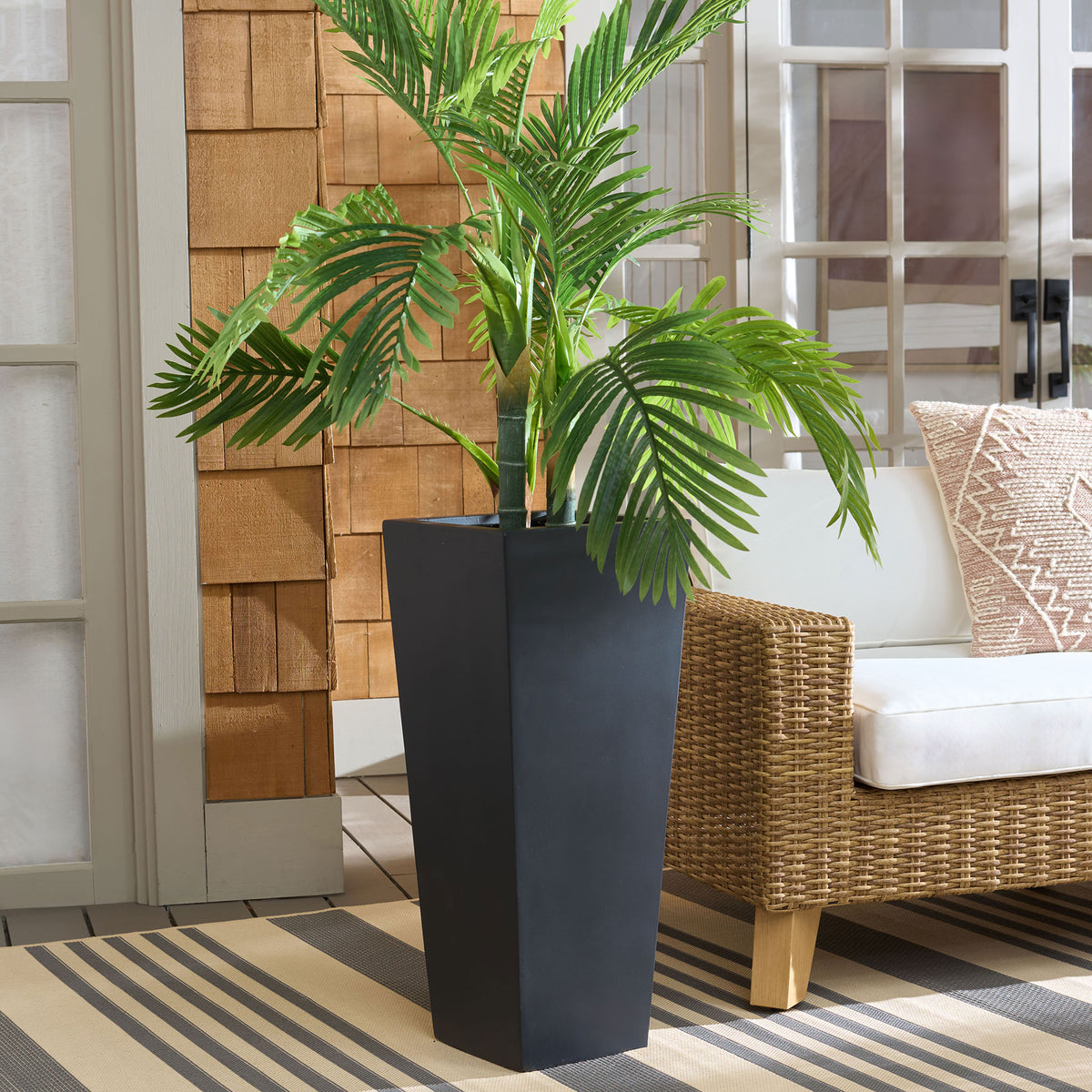 A tall black planter showcases lush palm leaves beside woven patio seating. || PAT1543A || front_angle_room