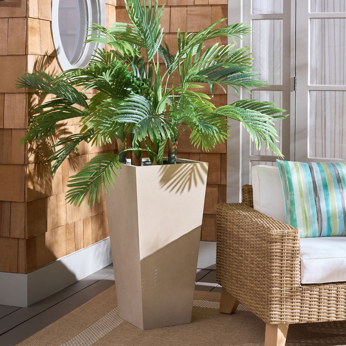 A large beige planter anchors lush palm foliage beside a woven outdoor chair. || PAT1544B || front_angle_room