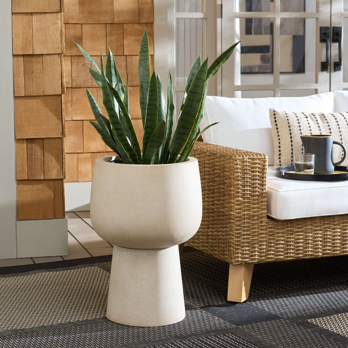 Beige pedestal planter holds leafy greenery beside a wicker patio chair || PAT1545B || front_room