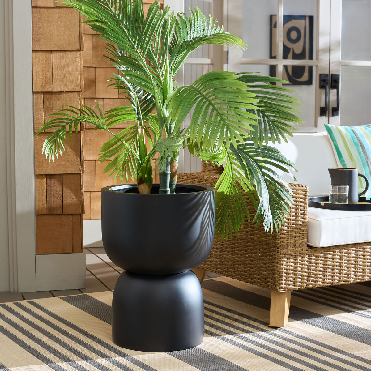 Black pedestal planter displays lush palm foliage beside woven seating. || PAT1546A || front_angle_room