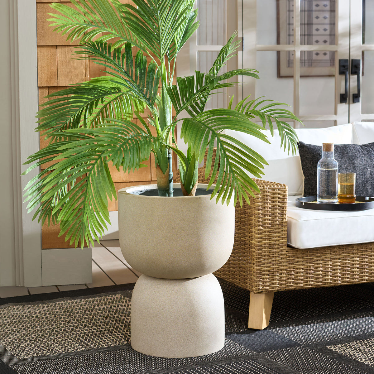 Beige pedestal planter holds lush palm foliage beside a woven lounge chair.