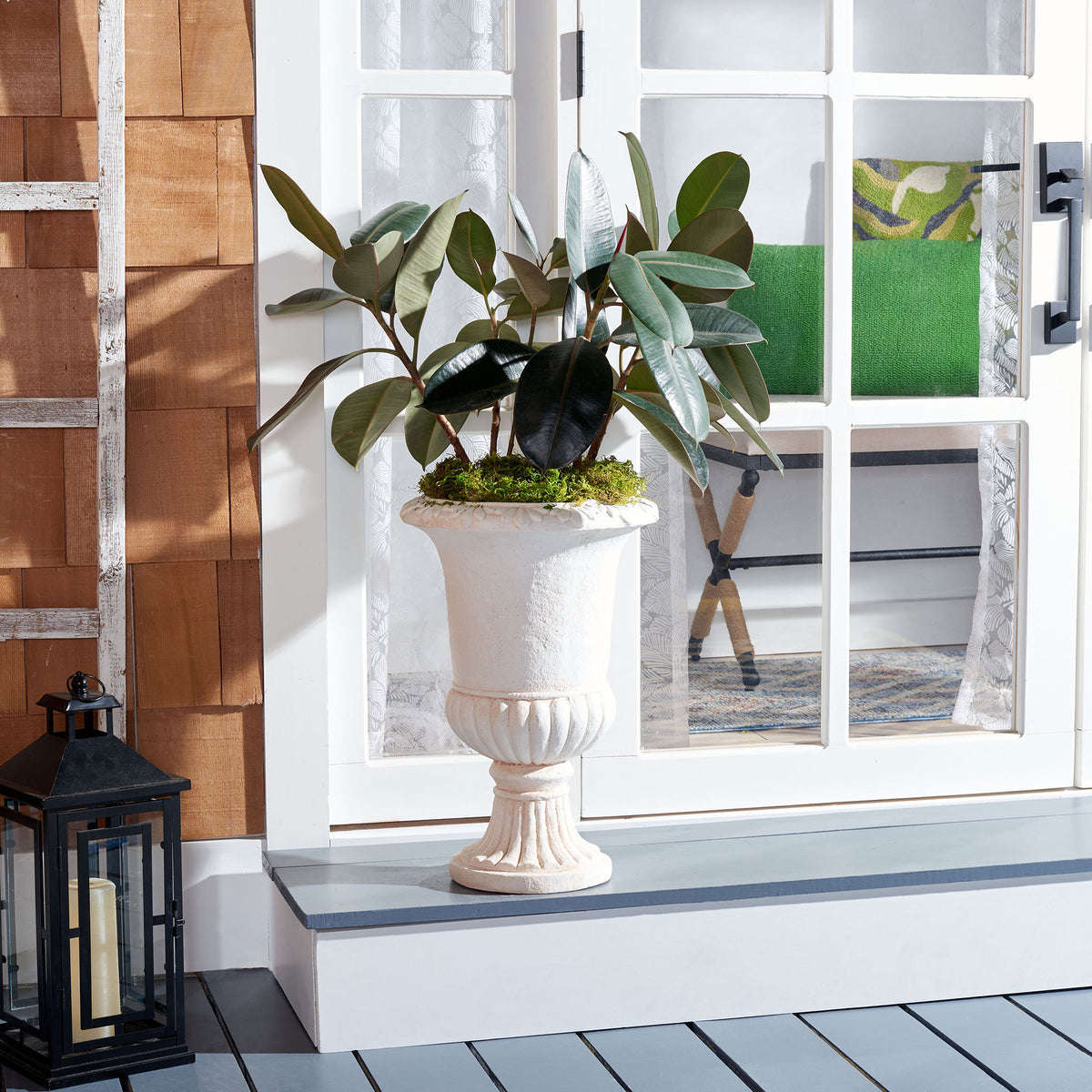 Off-white urn planter supports lush greenery against a bright doorway and warm wood siding.
