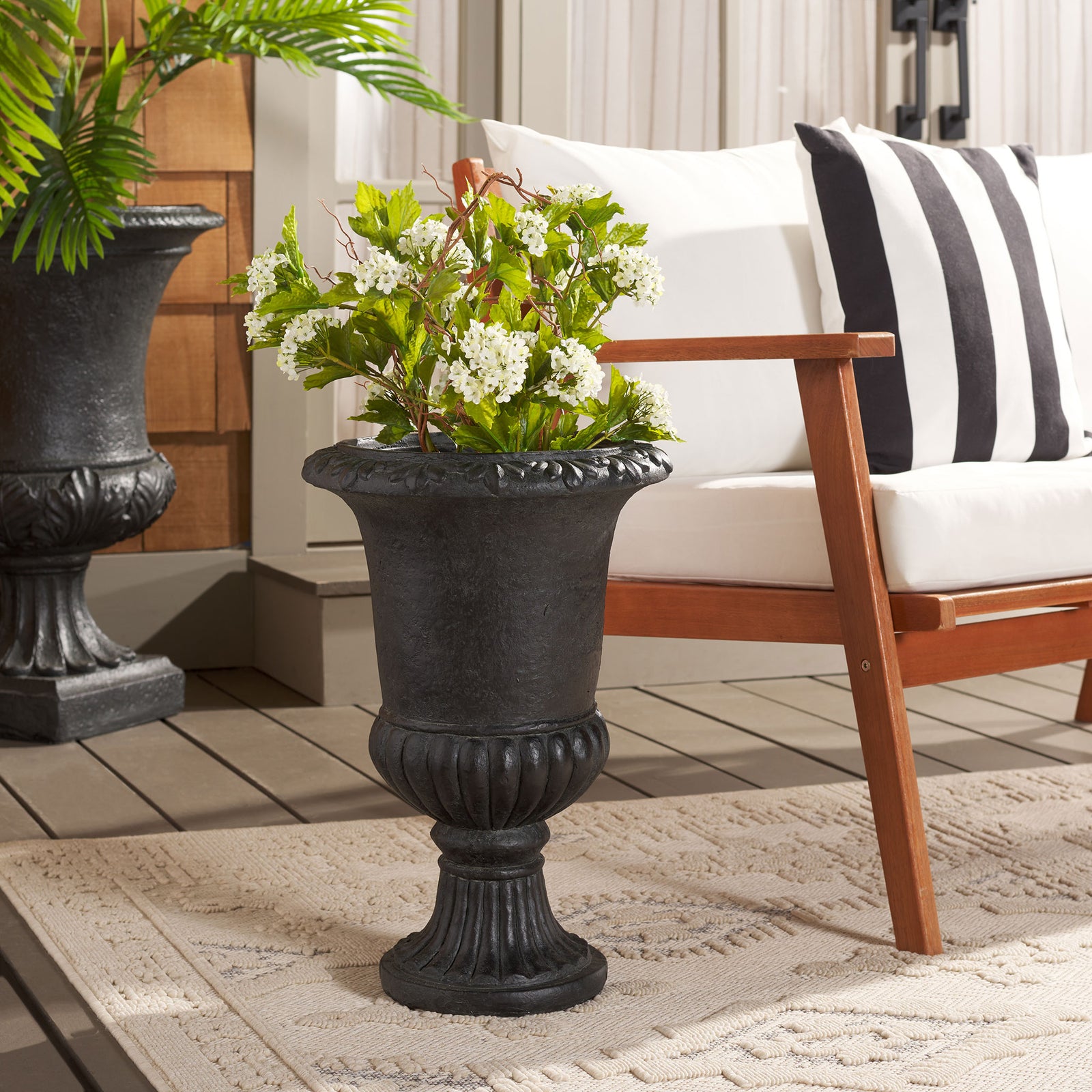 Black urn planter holds white blossoms and greenery beside a wood-framed chair. || PAT5037E || front_room