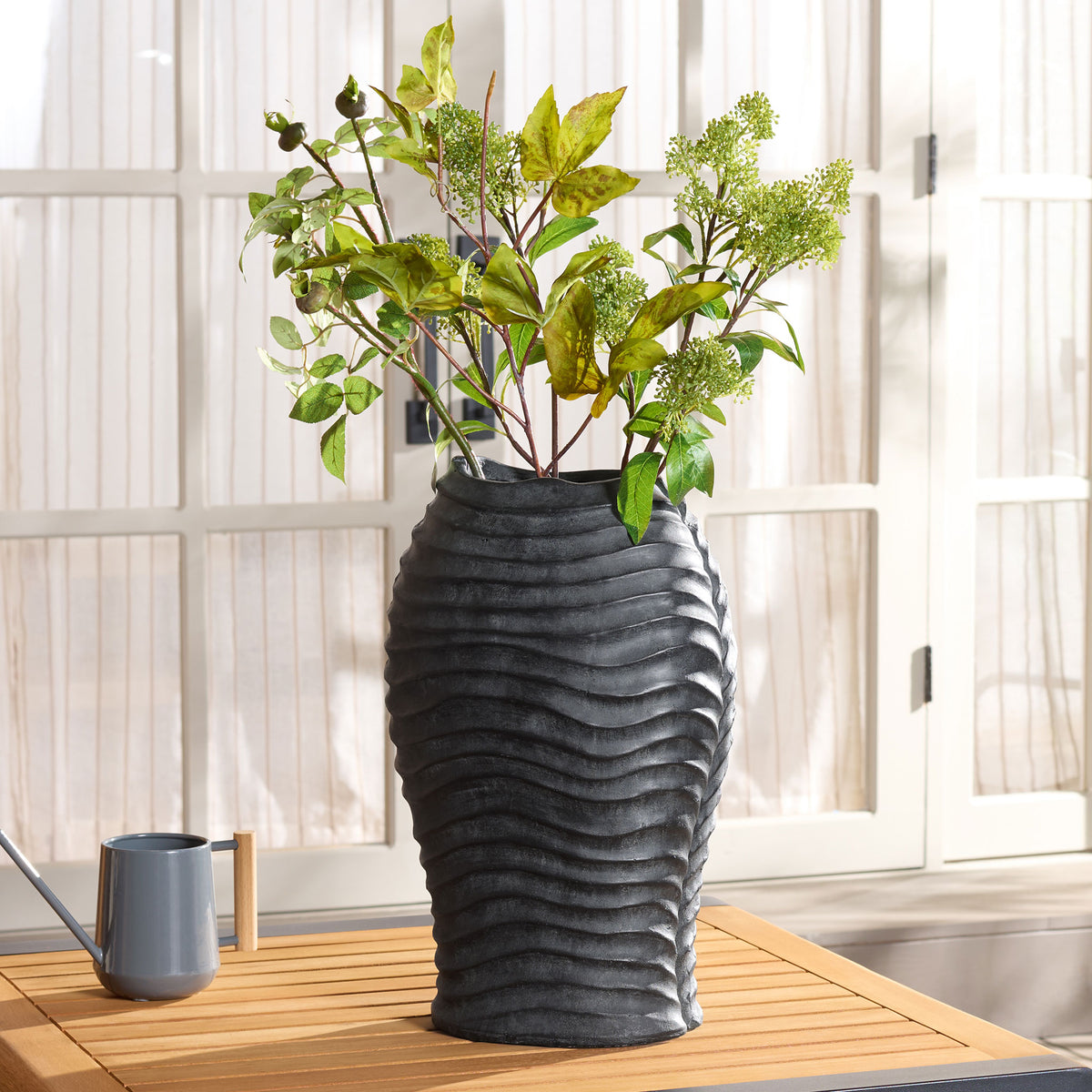 Leafy greenery fills a ribbed black planter on a sunlit slatted tabletop