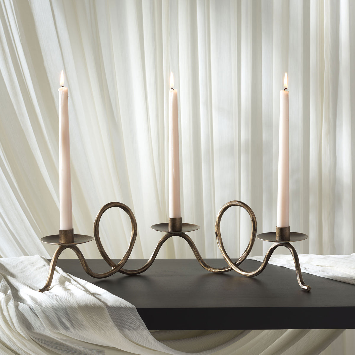 Three glowing taper candles crown an airy, wave-shaped metal centerpiece in a warm brass tone