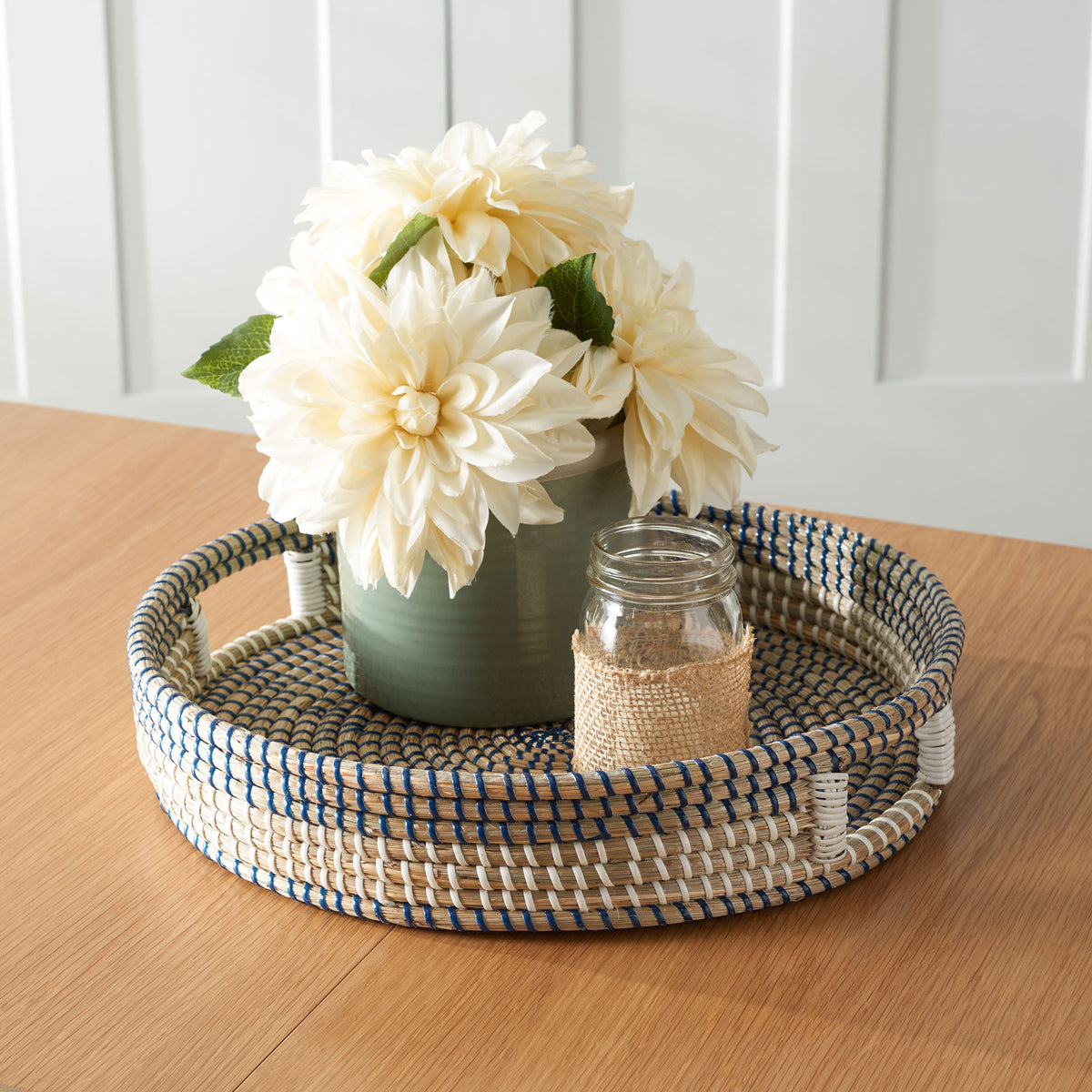 A blue-and-white woven tray holds a flower-filled vase and a small glass jar on a wood table. || STG1809A || front_angle_room