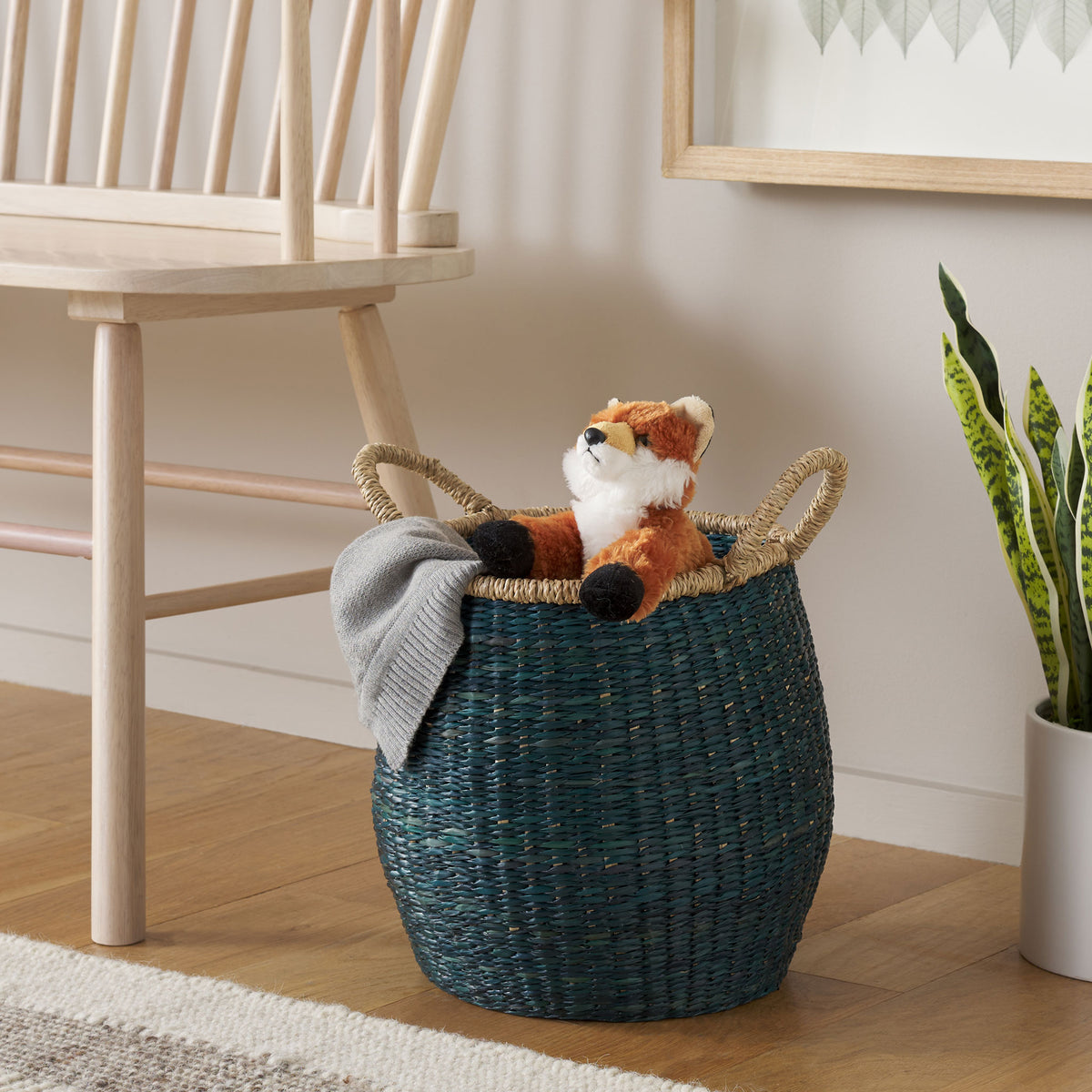 A green seagrass basket holds a cozy throw and plush toy beside a light wood chair || STG1815A || front_angle_room