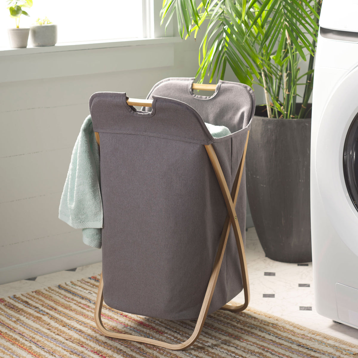 The gray fabric hamper holds laundry beside the washer on its natural bamboo frame. || STG1906A || front_angle_room