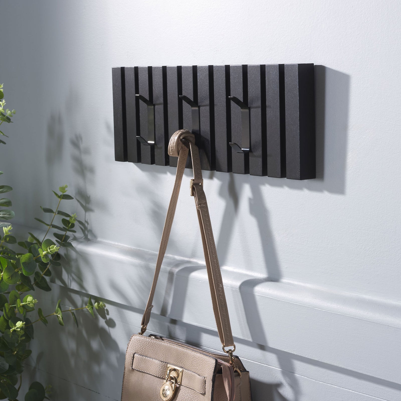 A beige handbag hangs from the middle hook of a black slatted wall rack built for everyday essentials. || STG5704E || front_angle_room