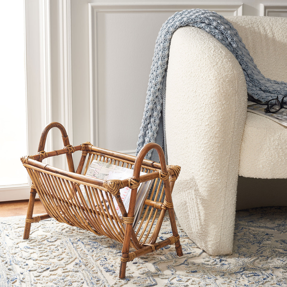 A honey-brown rattan newspaper rack holds reading material beside an upholstered chair || STG6500A || front_angle_room