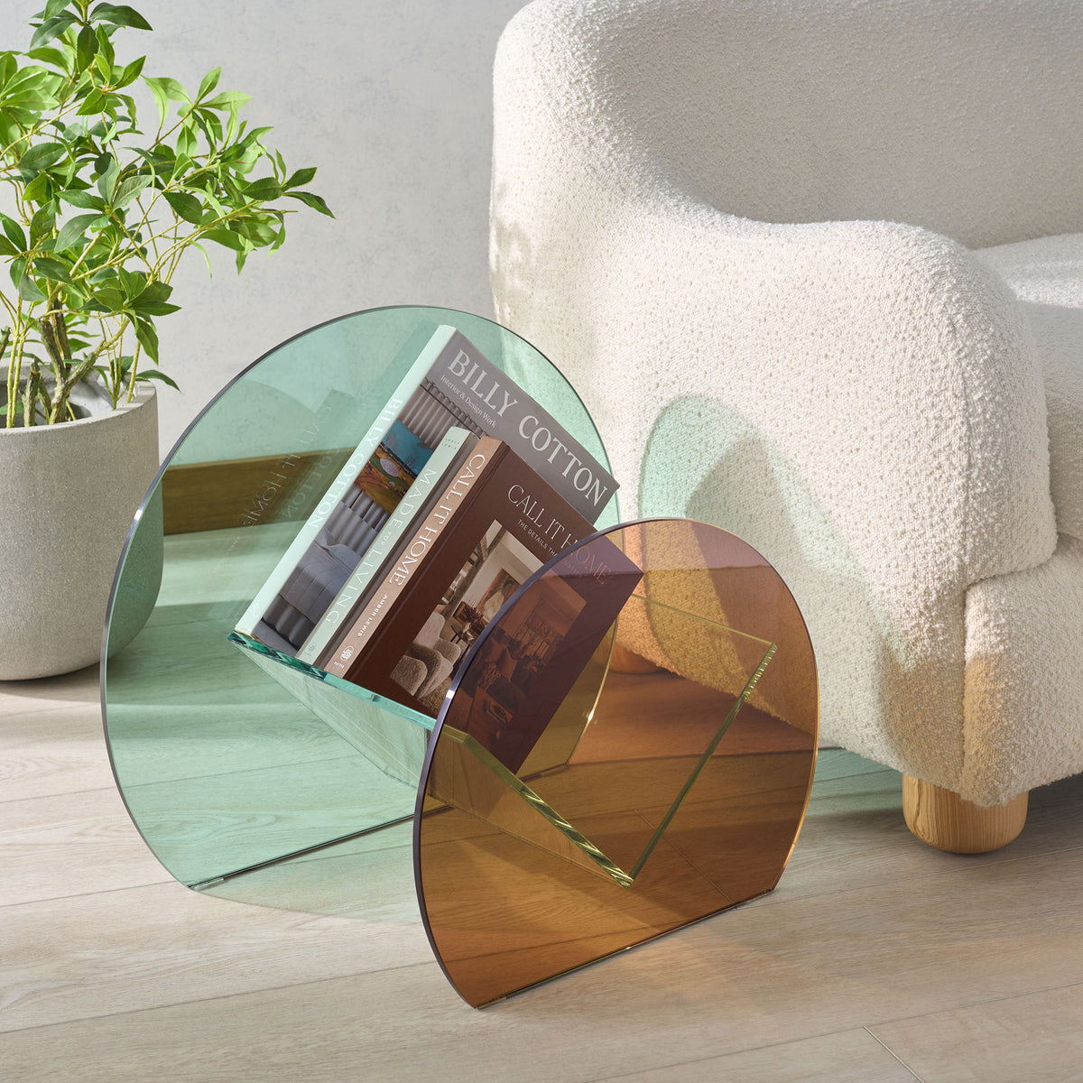 A clear glass cradle holds books between pale green and amber panels beside a cozy chair.