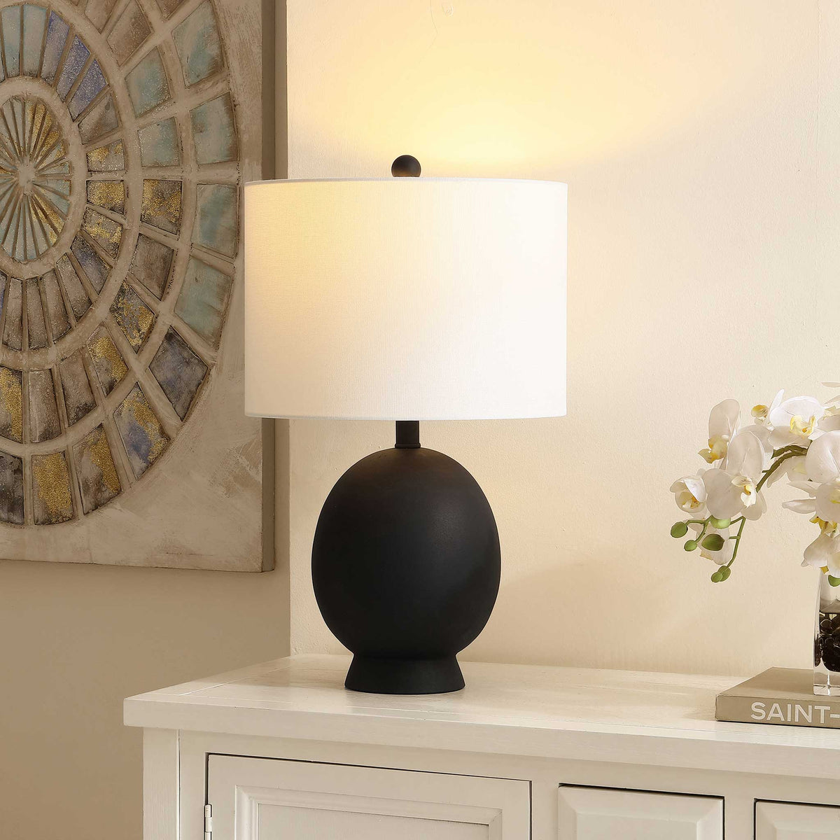 Soft, warm glow from a white drum shade over a matte black base creates cozy mood lighting in the room. || TBL4487A || front_angle_on_room