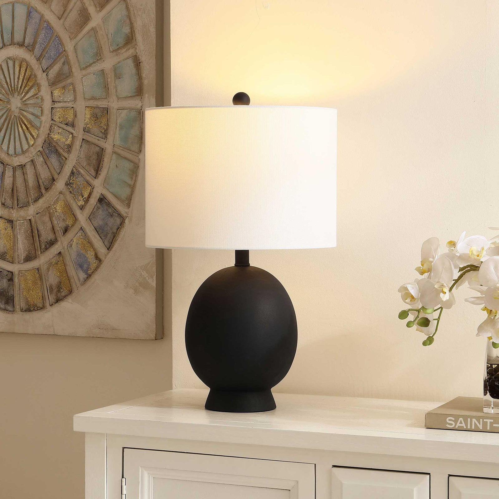 Soft, warm glow from a white drum shade over a matte black base creates cozy mood lighting in the room. || TBL4487A || front_angle_on_room