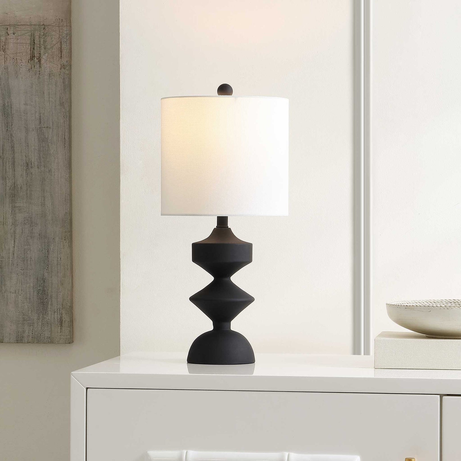 Warmly lit white drum shade atop a bold geometric black base for modern ambiance || TBL4501A || front_angle_on_room