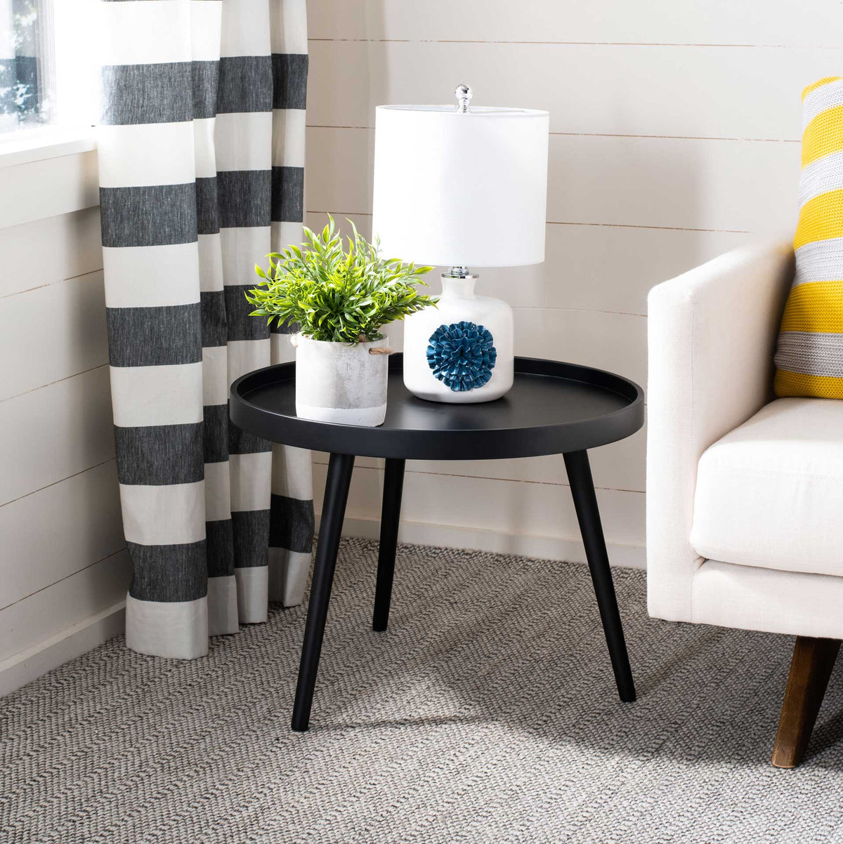 A round black tray table holds a lamp and greenery beside a white sofa.