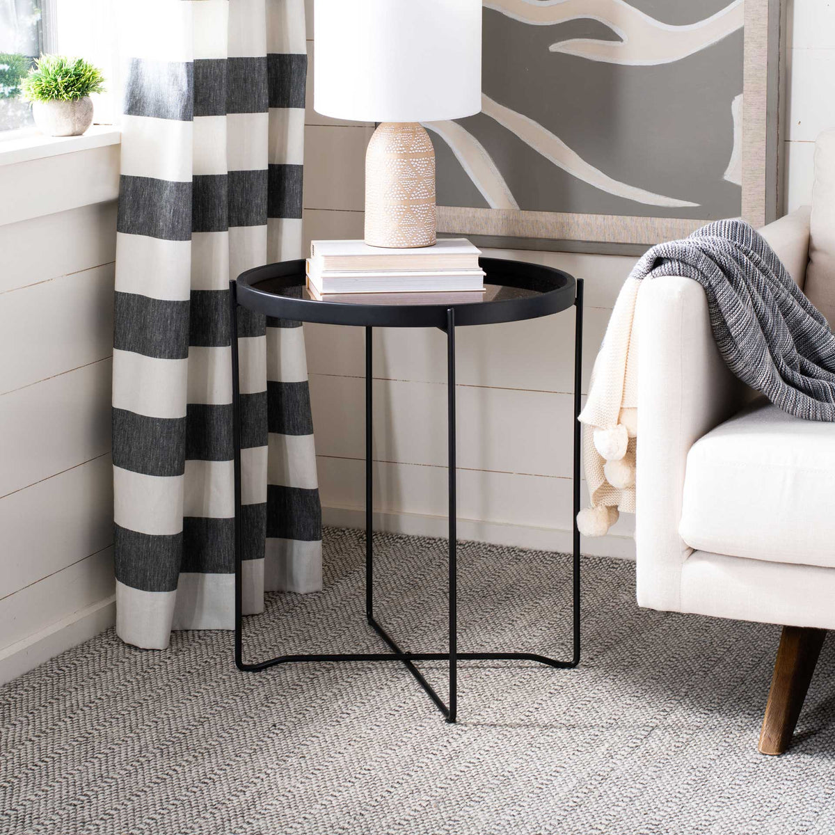 A slender black iron base supports a raised round tray that holds a lamp and books beside a sofa
