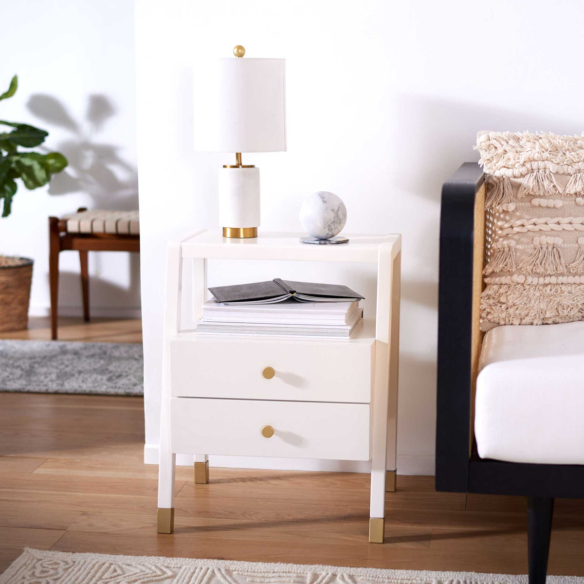 White bedside table holds reading materials on its open shelf, with two drawers and subtle gold accents