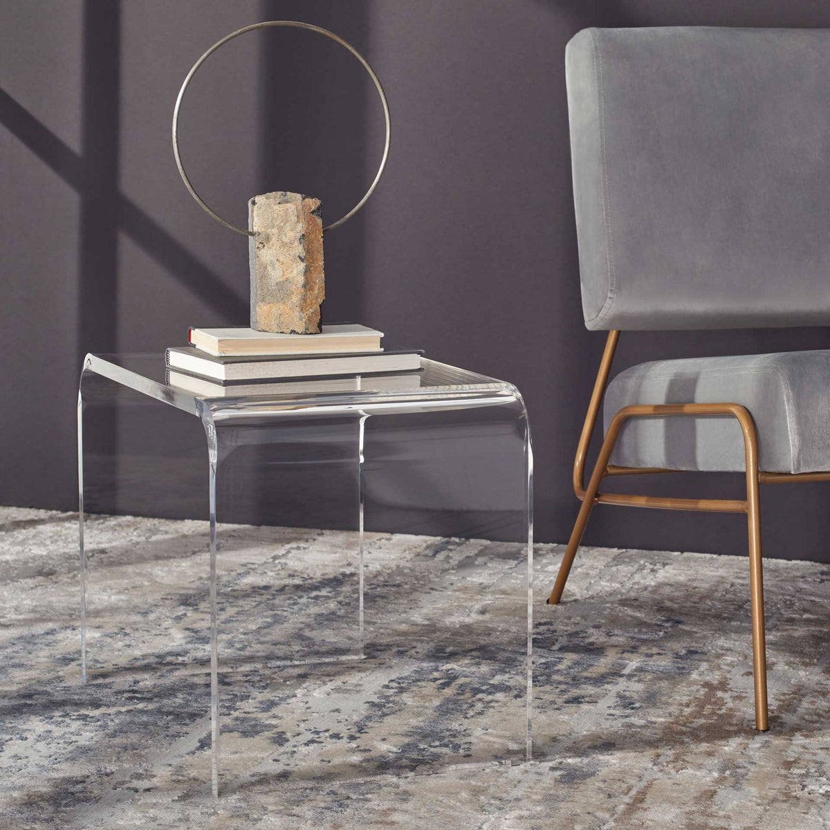 Books rest on a transparent acrylic side table beside a gray chair, giving the room an airy feel