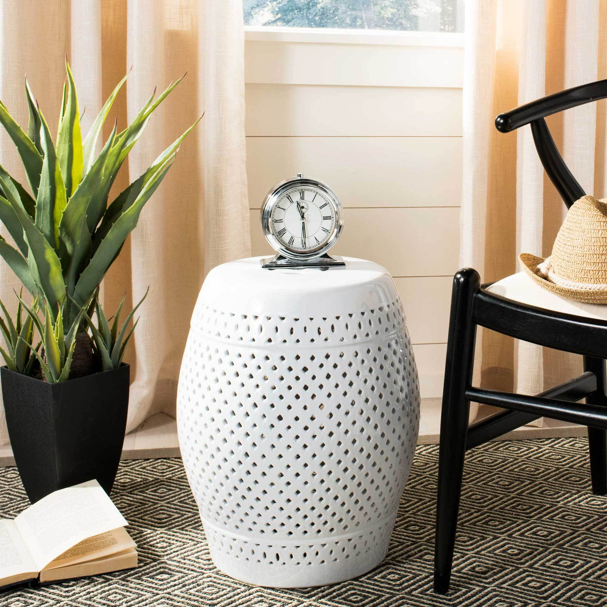Perforated ceramic stool holds a clock beside a black chair and leafy plant